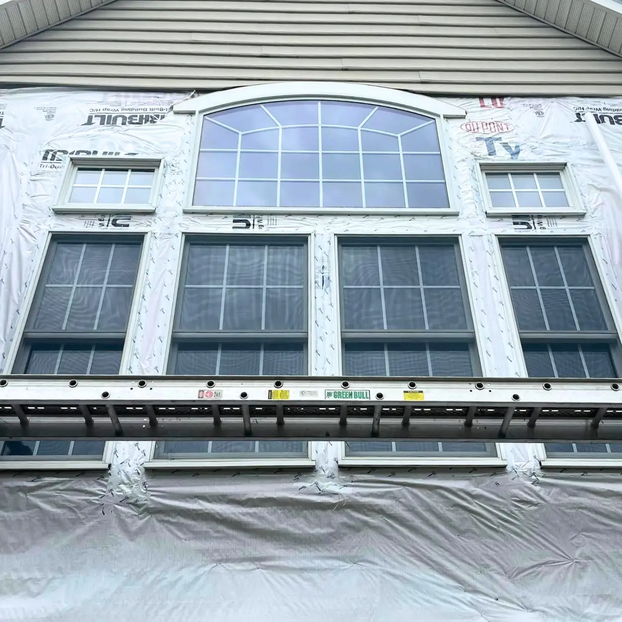 Two-story home during a vinyl siding replacement with new siding partially installed and underlayment visible.
